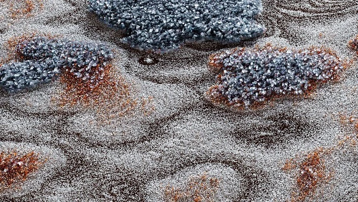 Close-up of a brown surface covered with clusters of silver and rust-colored pebbles, creating a textured, abstract, and glistening effect.