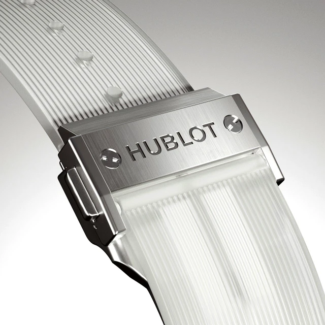 Close-up of a Hublot watch buckle and clear band showing the brand name against a neutral gradient background in a studio shot.