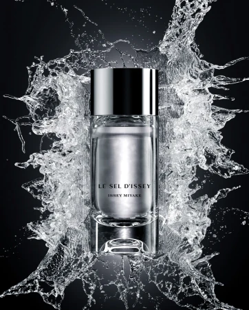 A cylindrical glass perfume bottle surrounded by a frozen splash of water against a dark black background.