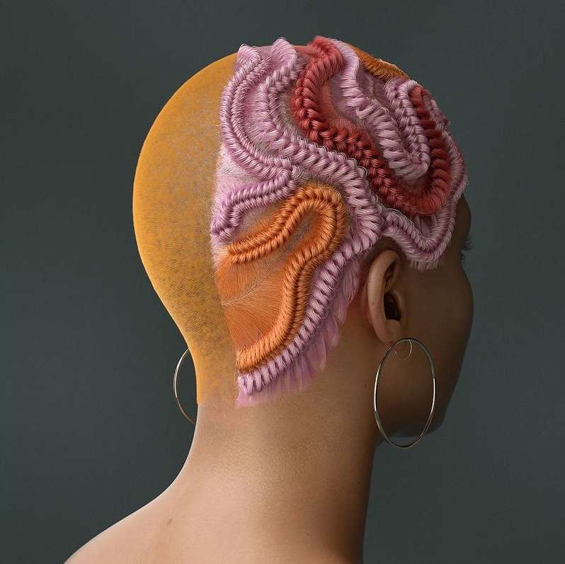 Artistic hairstyle with pink, red, and orange braids on a woman with a shaved head against a grey background; hoop earring visible.