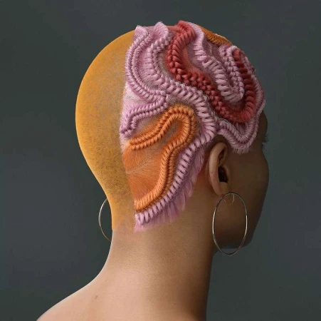 Artistic hairstyle with pink, red, and orange braids on a woman with a shaved head against a grey background; hoop earring visible.