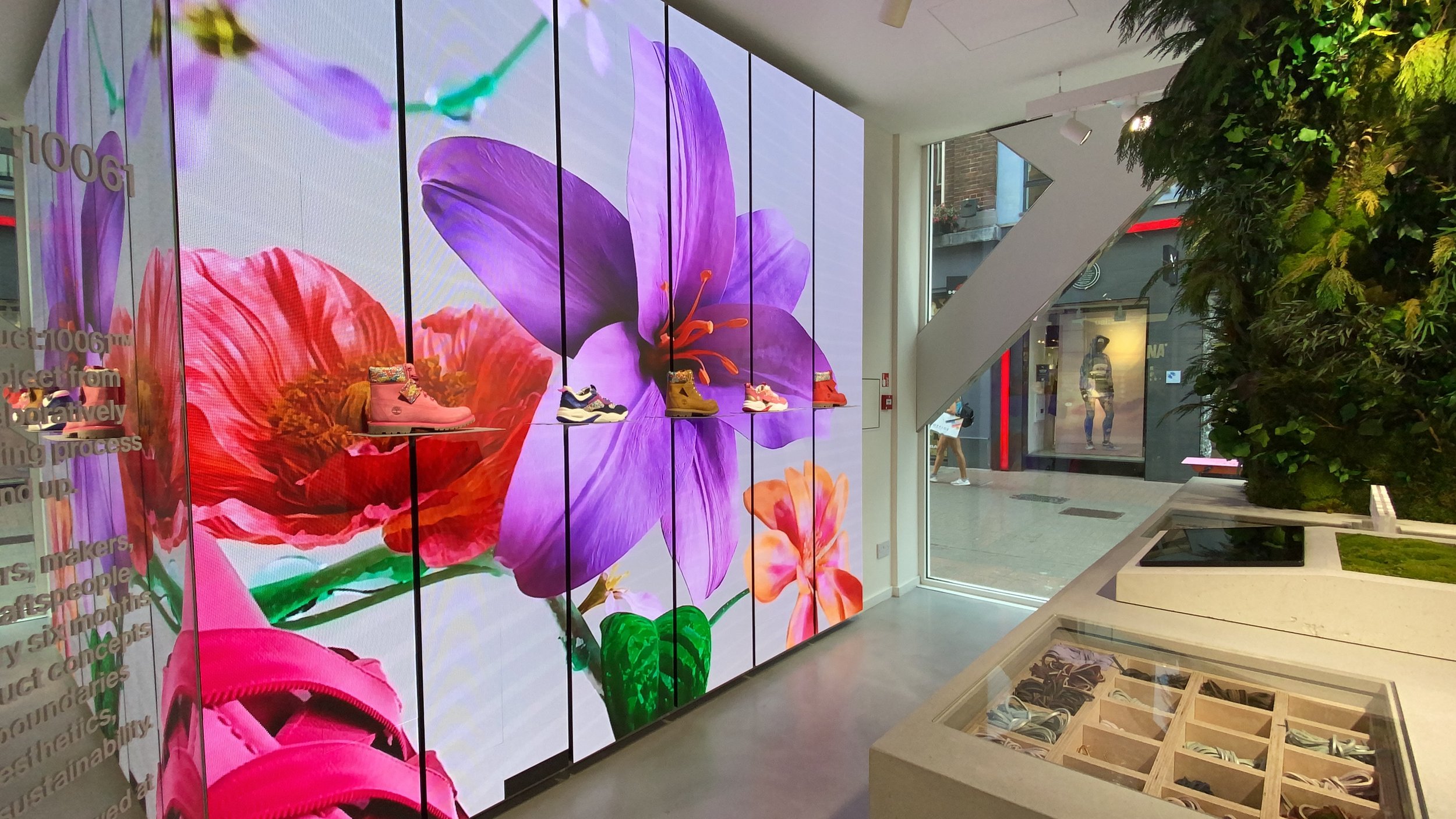 Retail store interior featuring a large digital screen wall displaying giant purple and red floral graphics with shoes on display shelves