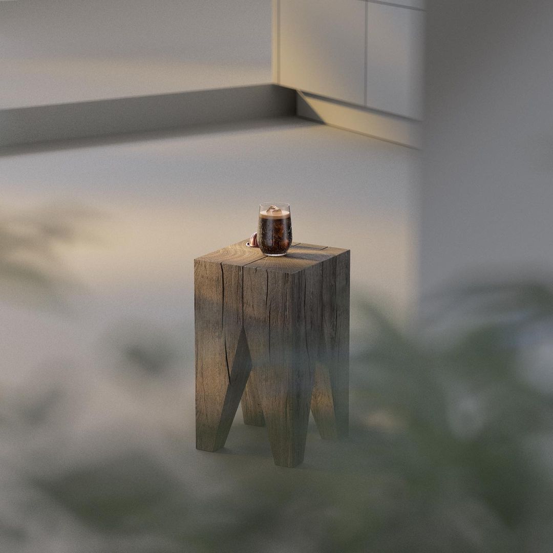 Iced coffee in glass sits atop a rustic wooden stool; the image's background features a blurred, minimalist interior and greenery.