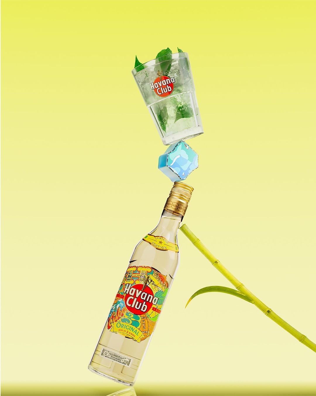Still-life shot of a bottle of Havana Club rum, a glass of mojito, an ice cube, and a sugarcane stalk against a gradient yellow background.