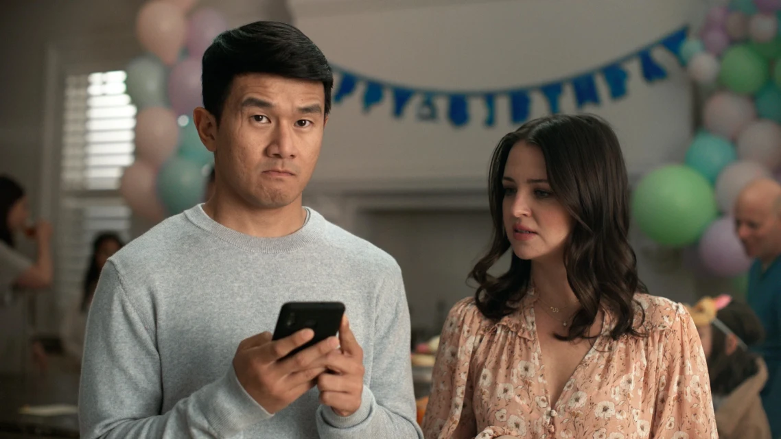 Adult man and woman pose indoors near birthday decorations. Man holds a cell phone with a concerned expression; woman looks on with concern.