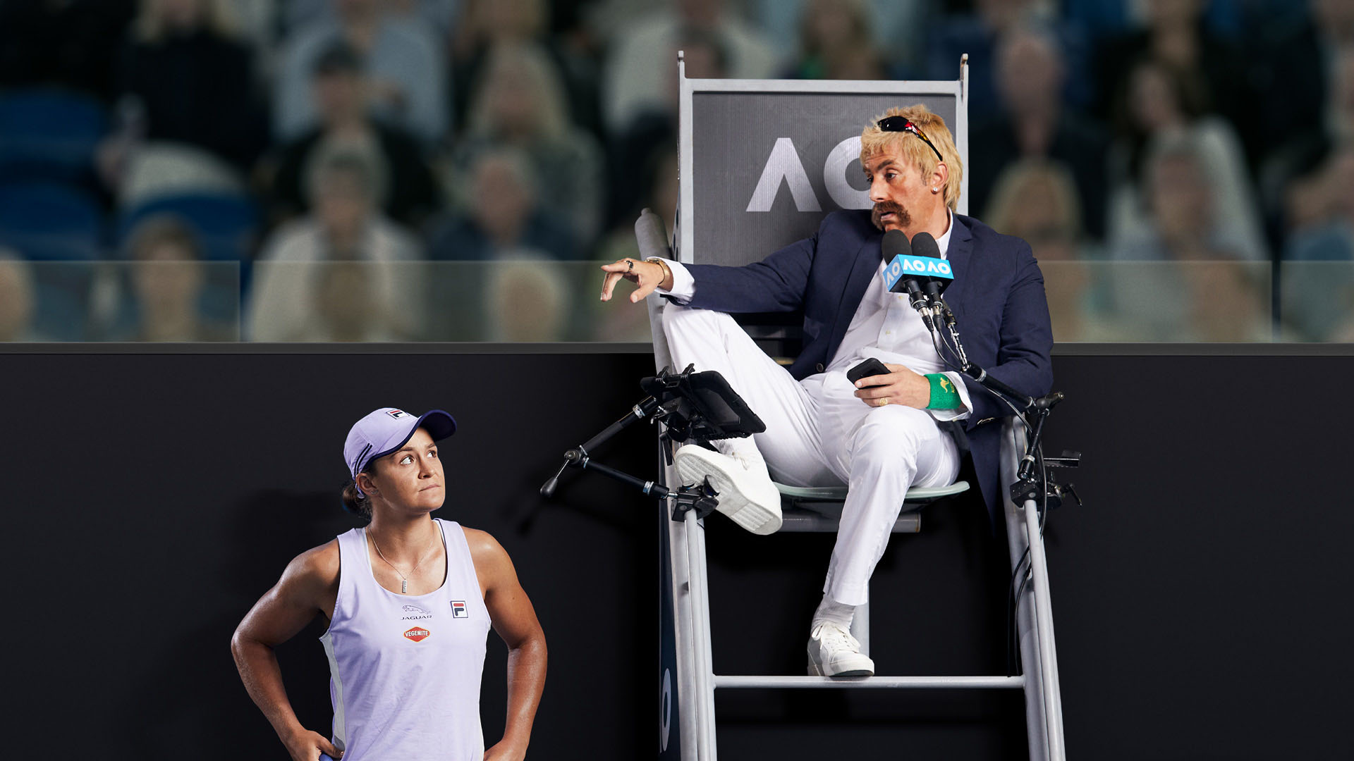 Tennis match official impersonating John McEnroe sits in an umpire chair, while a female tennis player looks up at him on the court.