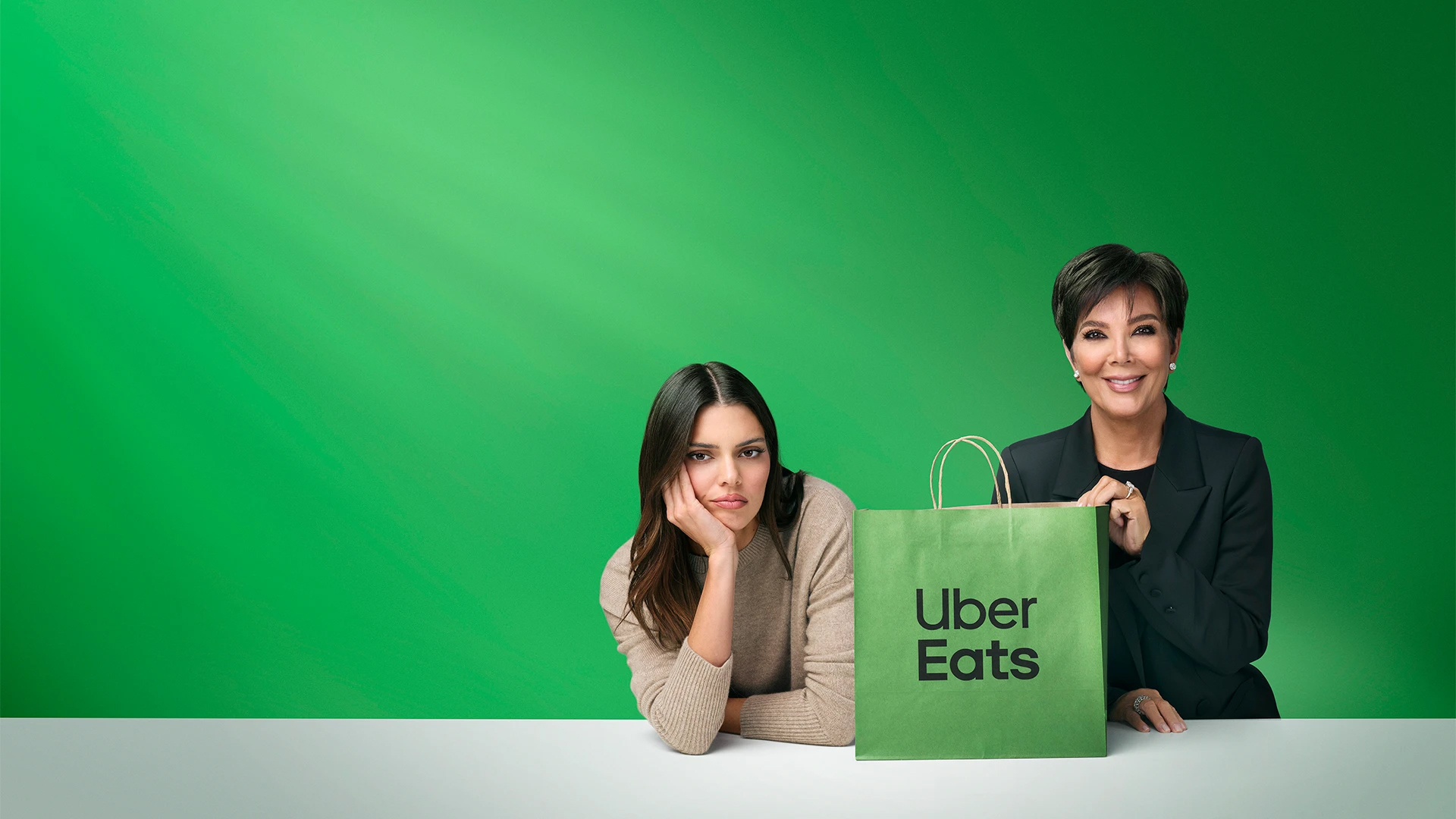 Two women, one looking glum, the other smiling, are posing with an Uber Eats delivery bag against a bright green background. 