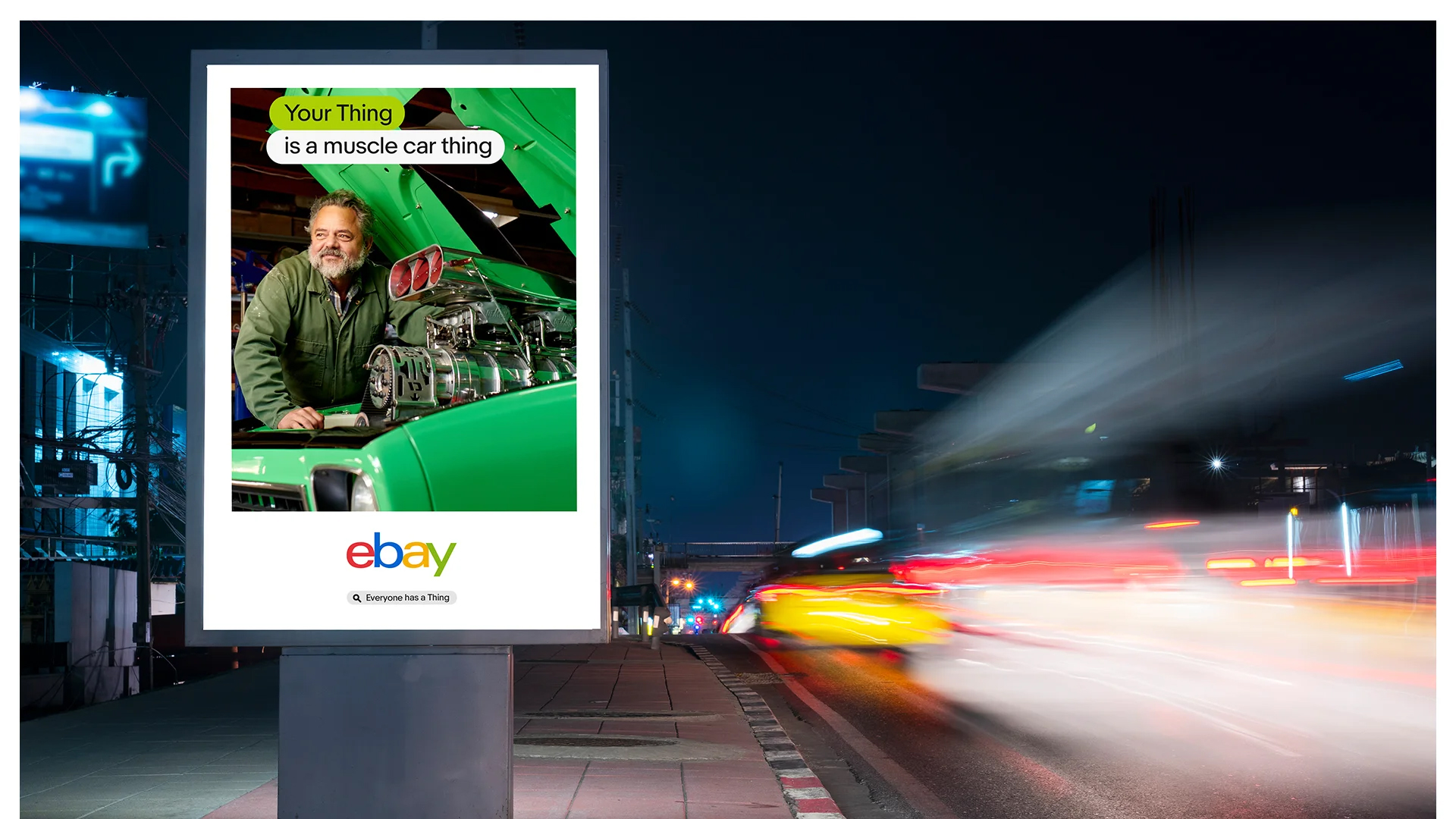 Ebay advertising billboard shows a man working on a green muscle car at night with colorful motion-blurred traffic background.