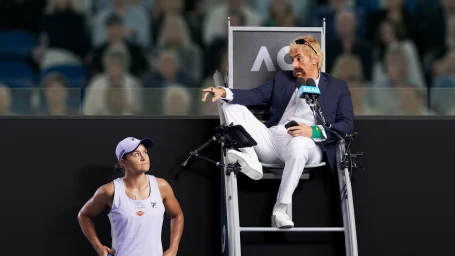 Tennis player in white sports apparel standing below an umpire chair with a man in white trousers and blazer gesturing.