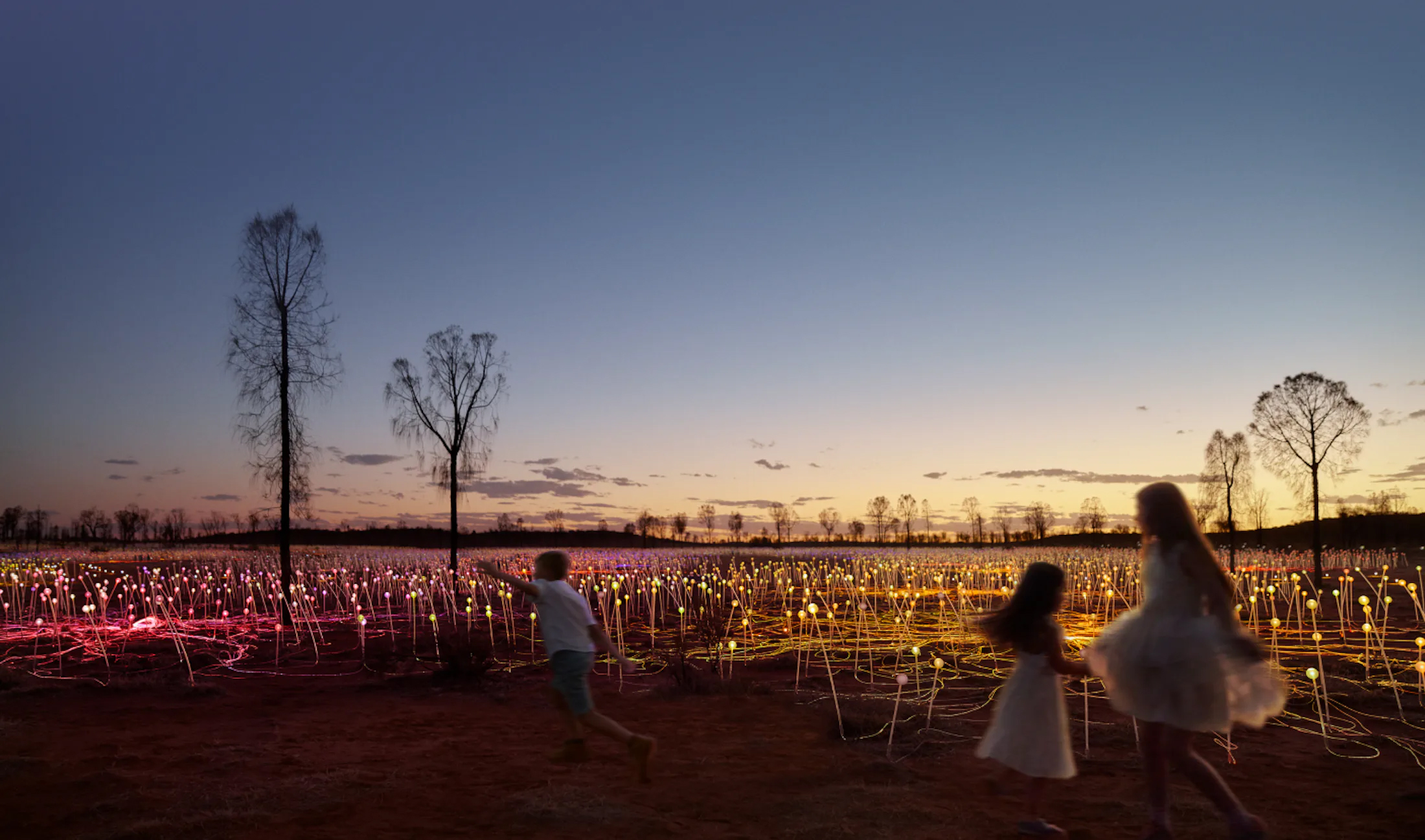 Three children, a boy and two girls, run through a field with decorative illuminated lights at dusk, with trees silhouetted in background.