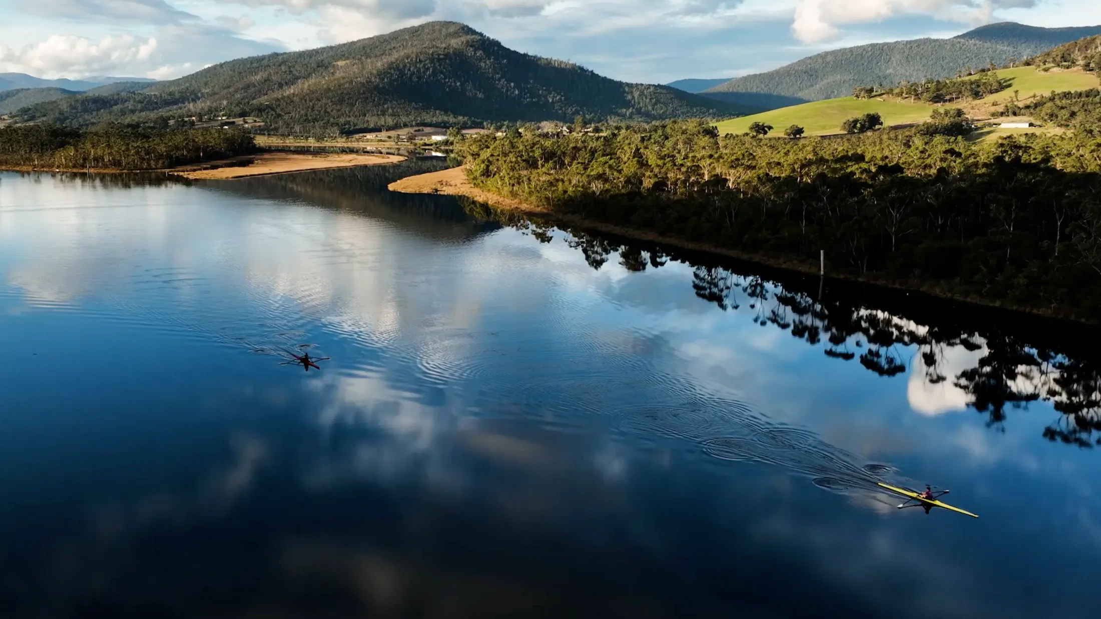 Aerial view of two rowboats gliding across a serene lake reflecting the sky, with lush green hills and mountains in the backdrop.