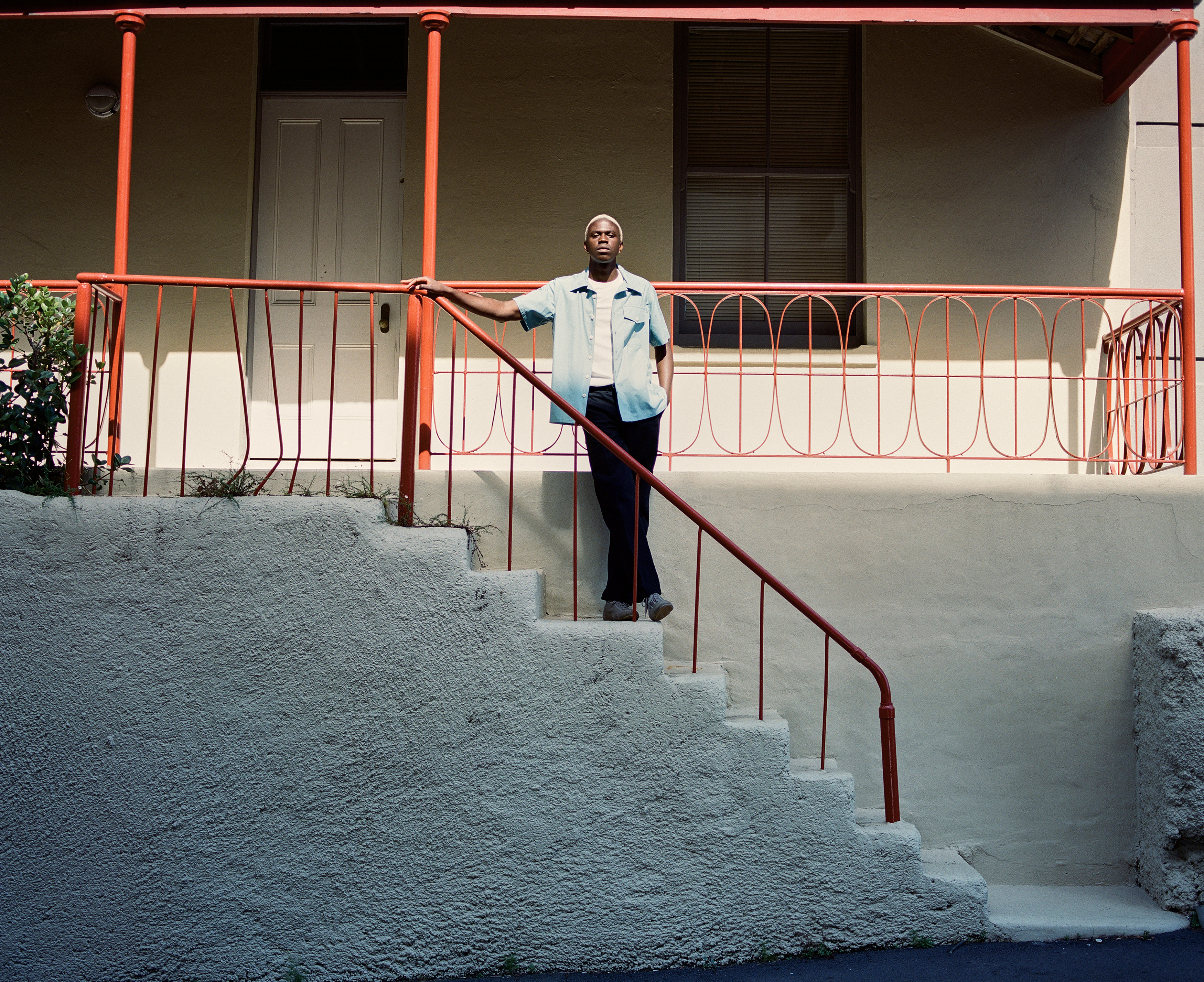 Adult Black man in casual clothes posing on concrete stairs with a red railing leading up to a white stucco house with a closed door.
