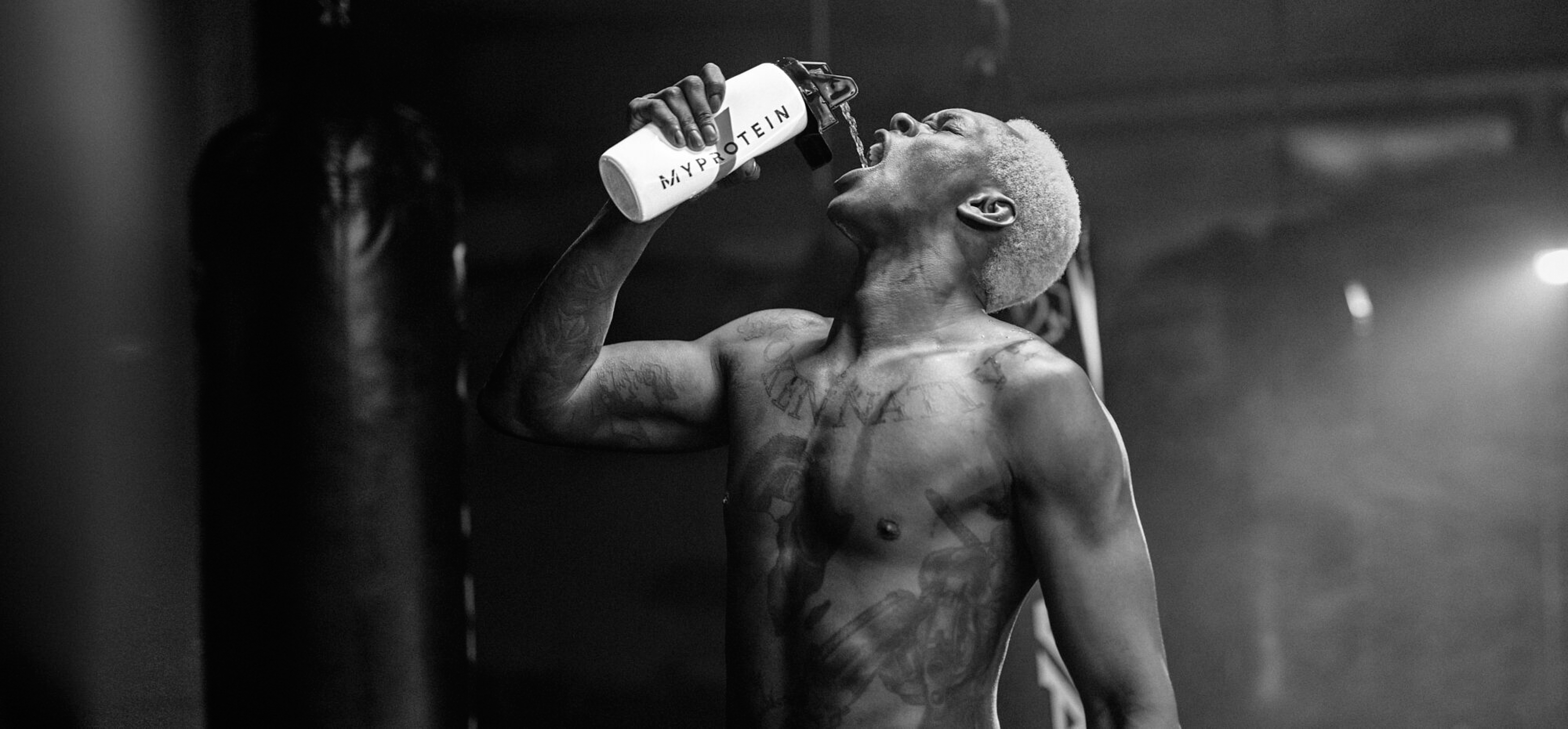 Muscular man drinking from a MyProtein water bottle in a dark gym with punching bags. Monochrome shot of the fitness workout.
