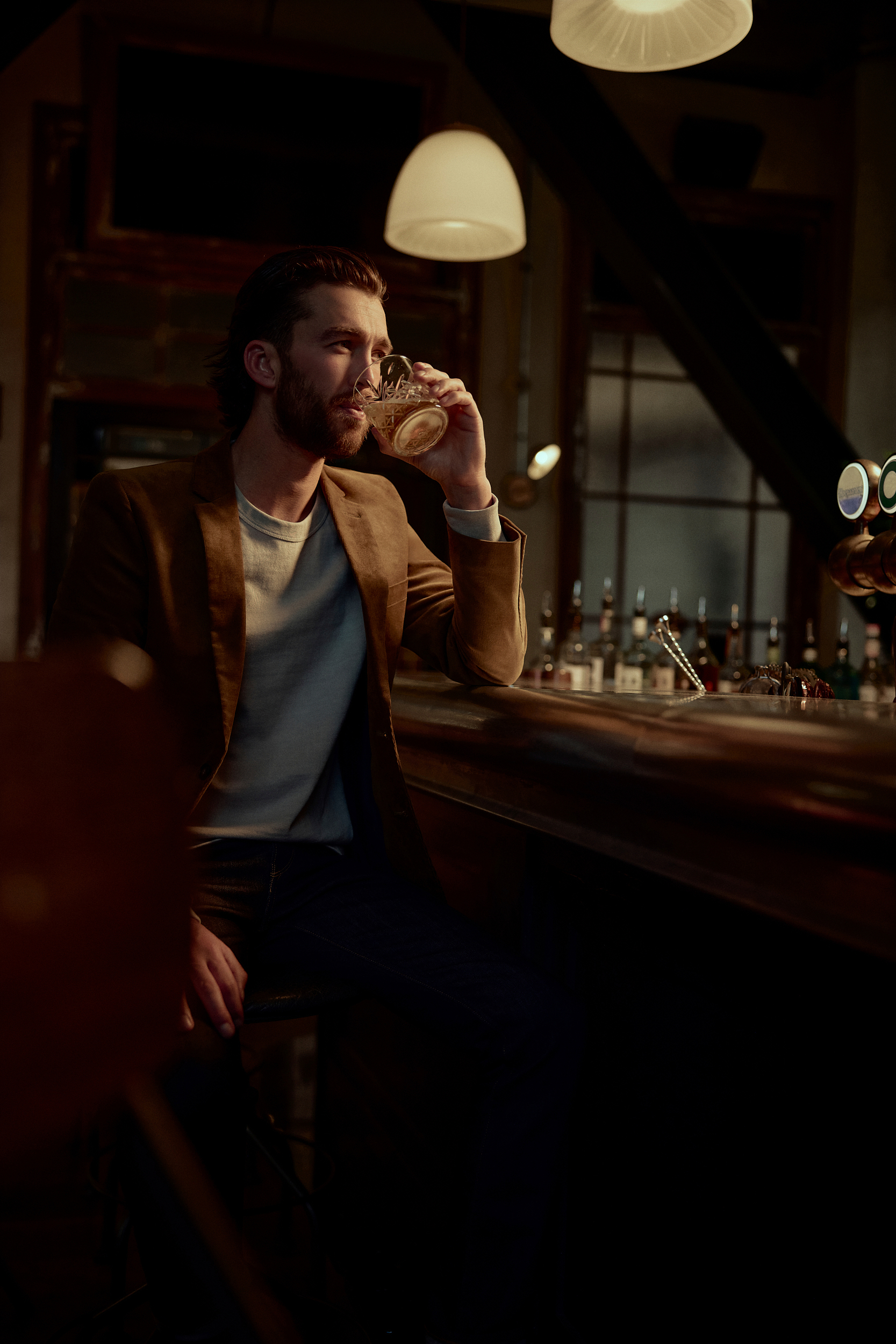 Man with a beard drinking from a glass at a bar, wearing a brown jacket and a light blue shirt, with bottles in the background.