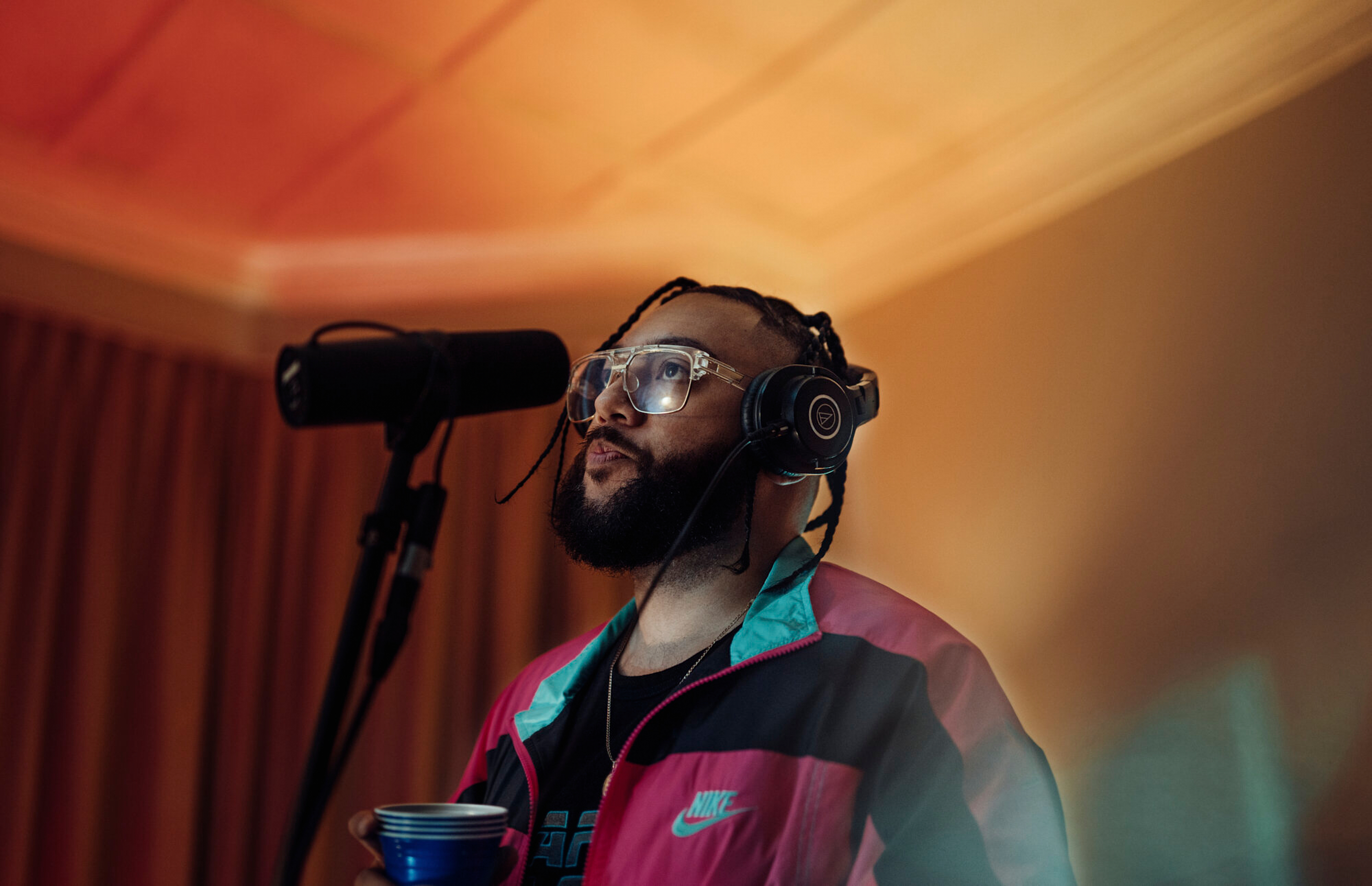 Man with beard and glasses wearing headphones and pink jacket in recording studio in front of a microphone with orange light.