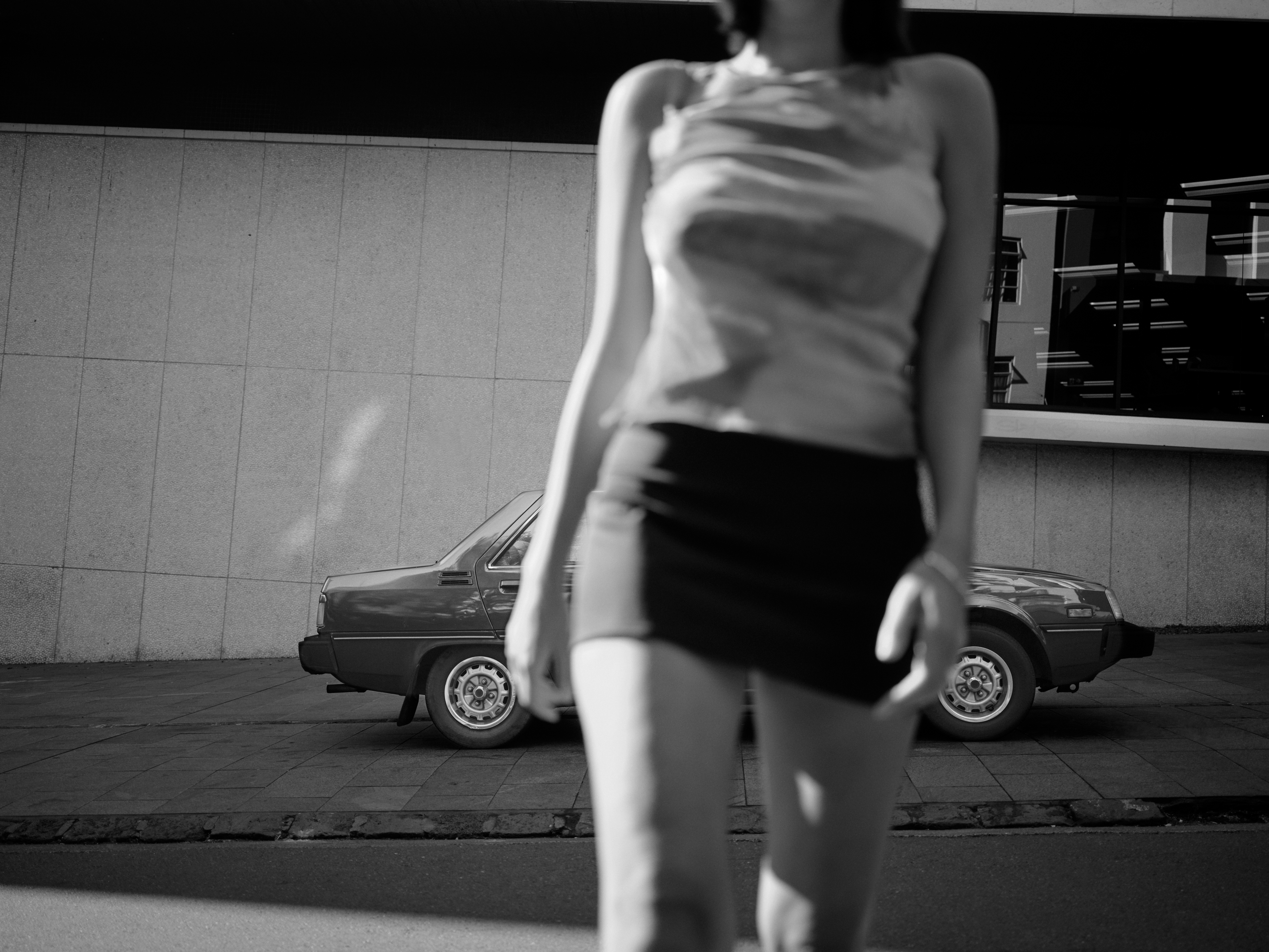 Black and white shot of an adult in a mini skirt standing in front of a vintage car, with a modern building in the background.