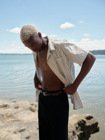 Young man with short blonde hair wearing an open striped short-sleeve shirt and dark trousers standing near the water.