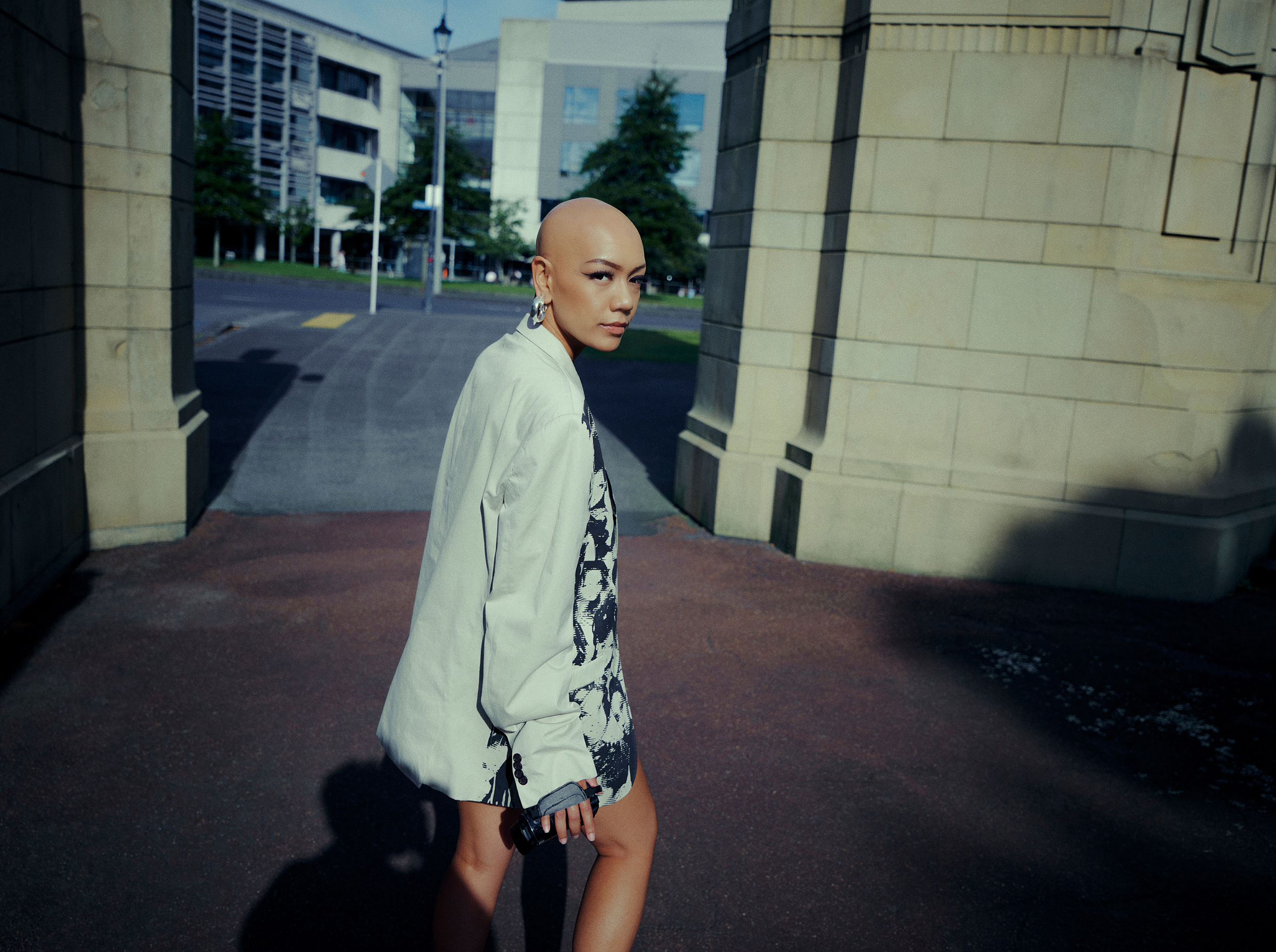 Stylish bald woman in patterned jacket walks through stone archway on a city street with modern architecture in background on a sunny day.