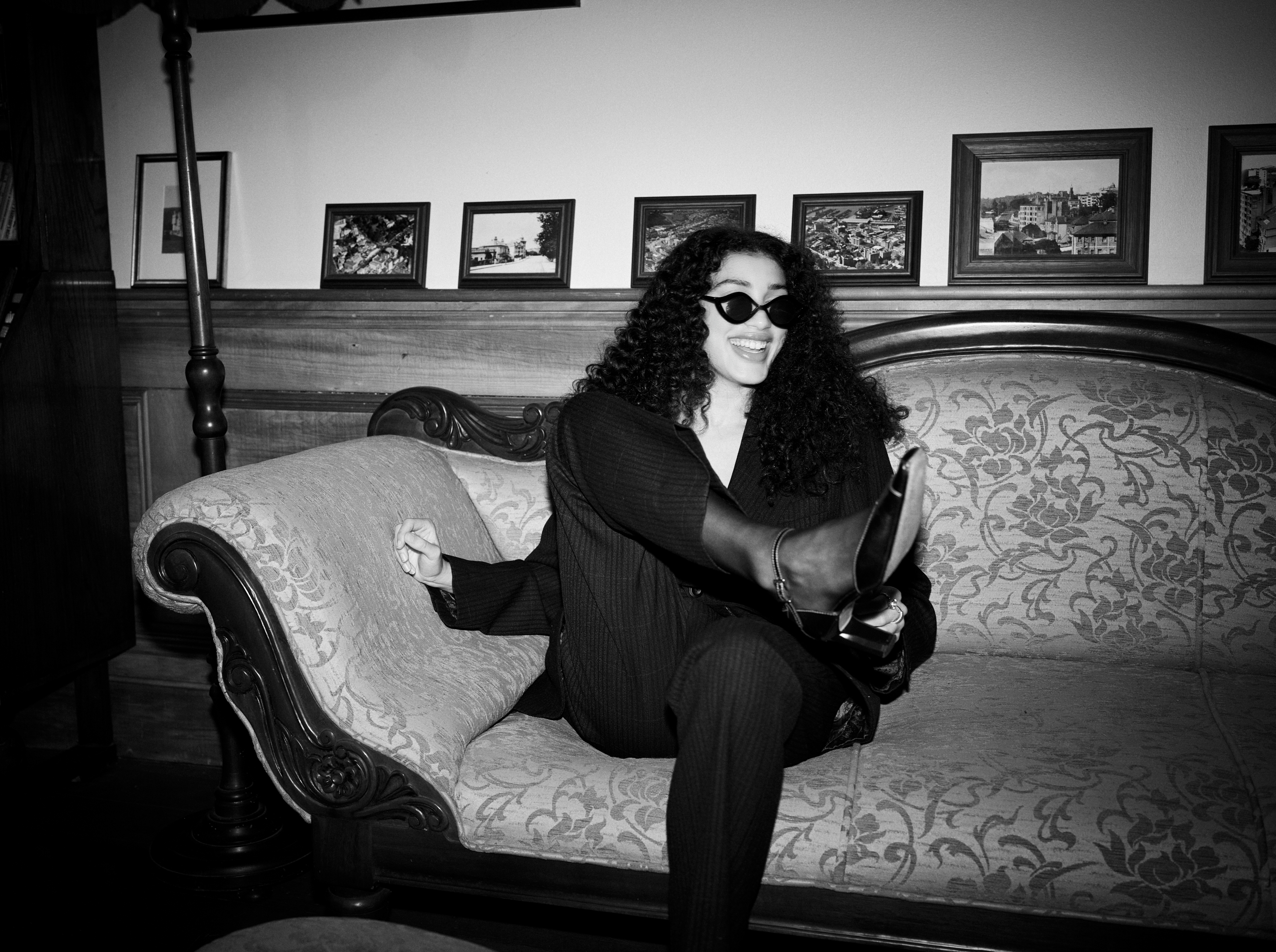 A woman with curly hair and sunglasses, wearing a dark suit, sits on a patterned vintage couch indoors with framed pictures on the wall.