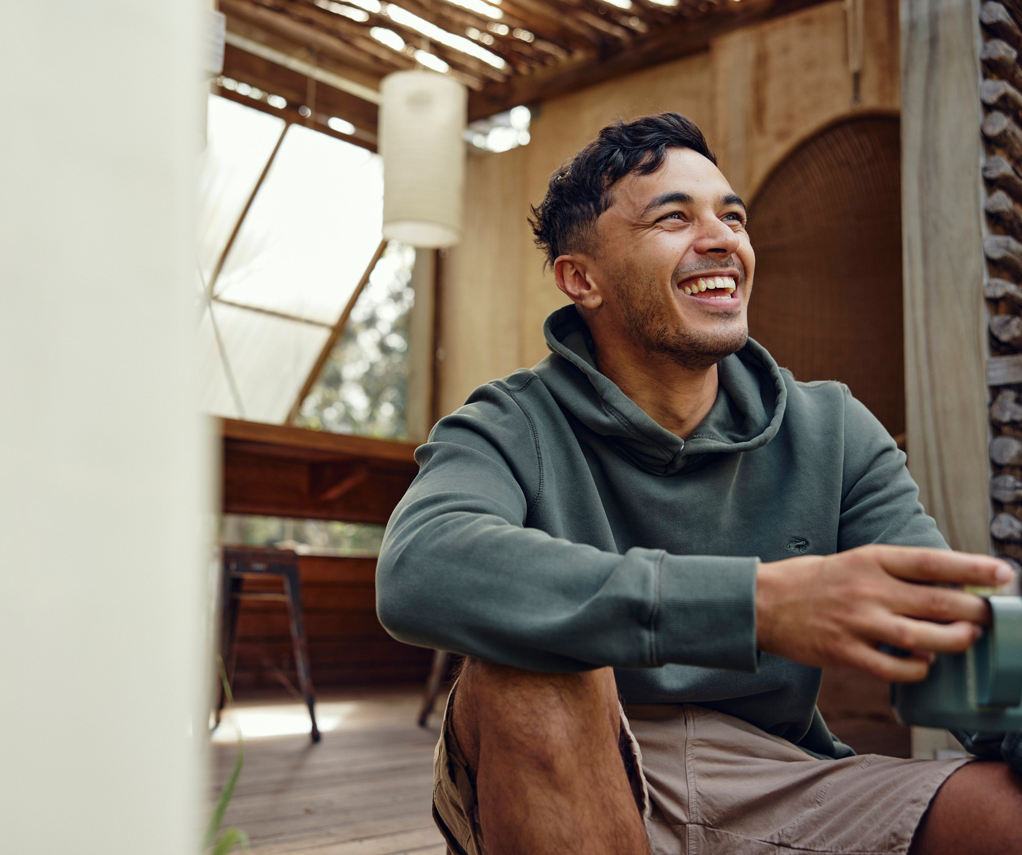 Man smiles in a green hoodie sitting cross-legged indoors, holding a cup with natural light and earthy tones. Relaxed lifestyle.