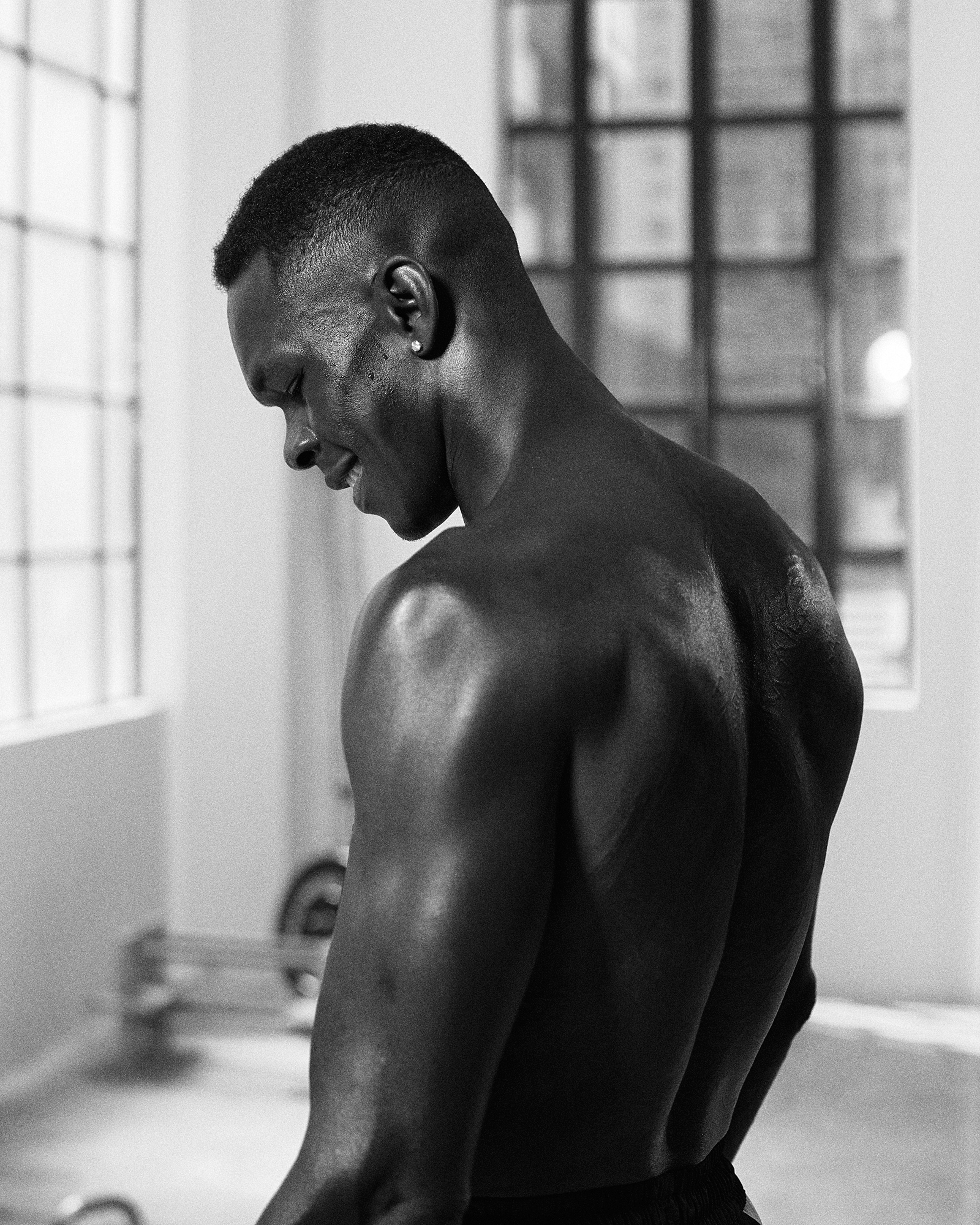 Monochromatic studio shot of a muscular, bare-chested African American man looking down, framed by a barbell and large windows.