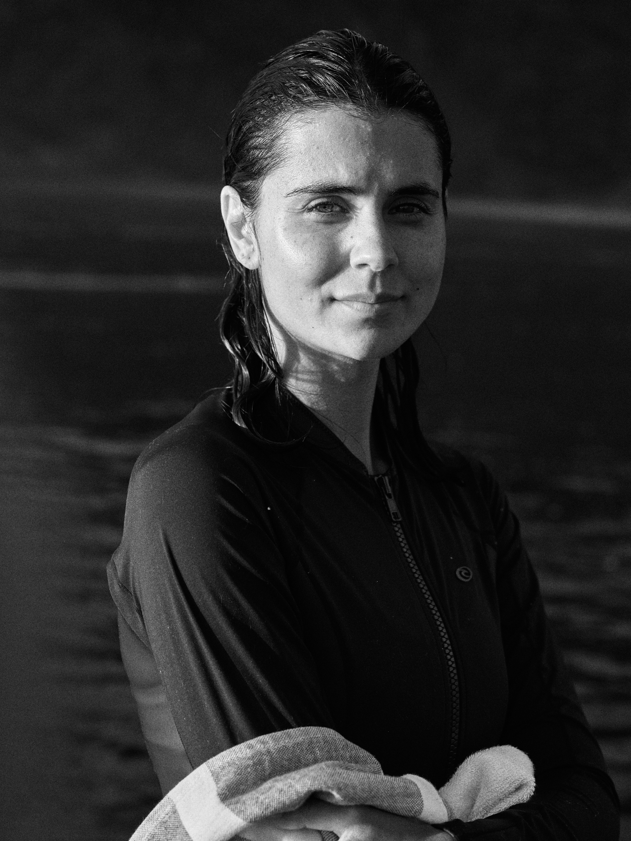 Black and white portrait of a woman with wet hair wearing a dark top, standing in front of dark water.