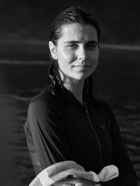 Black and white portrait of a woman with wet hair wearing a dark top, standing in front of dark water.