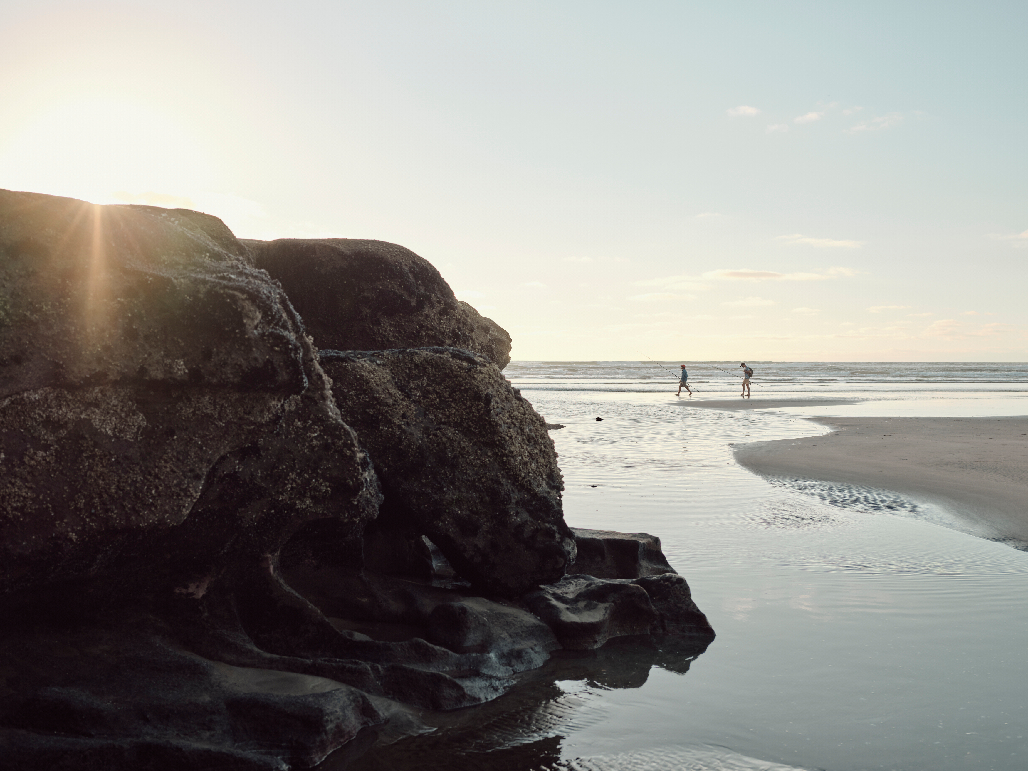 Scenic beach landscape with rock formations and two people walking in the distance along the water's edge at sunset.