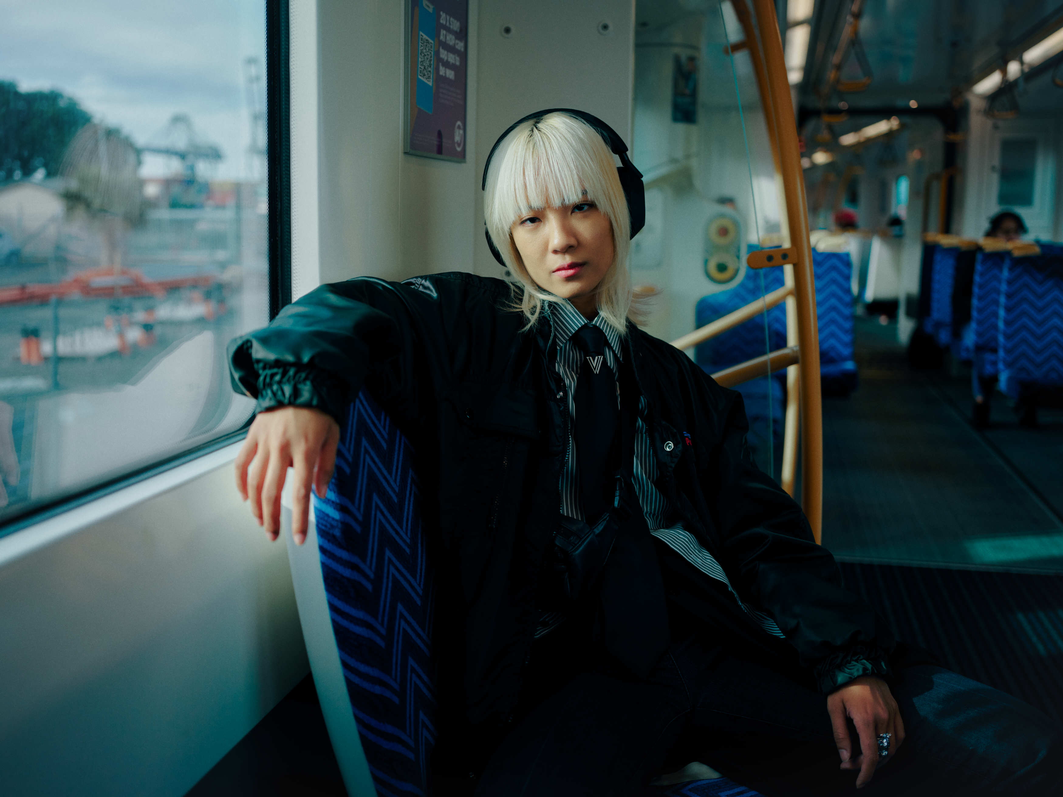 Young adult with striking platinum blonde hair sits on a train, wearing a black jacket, striped shirt and tie, gazing intently.