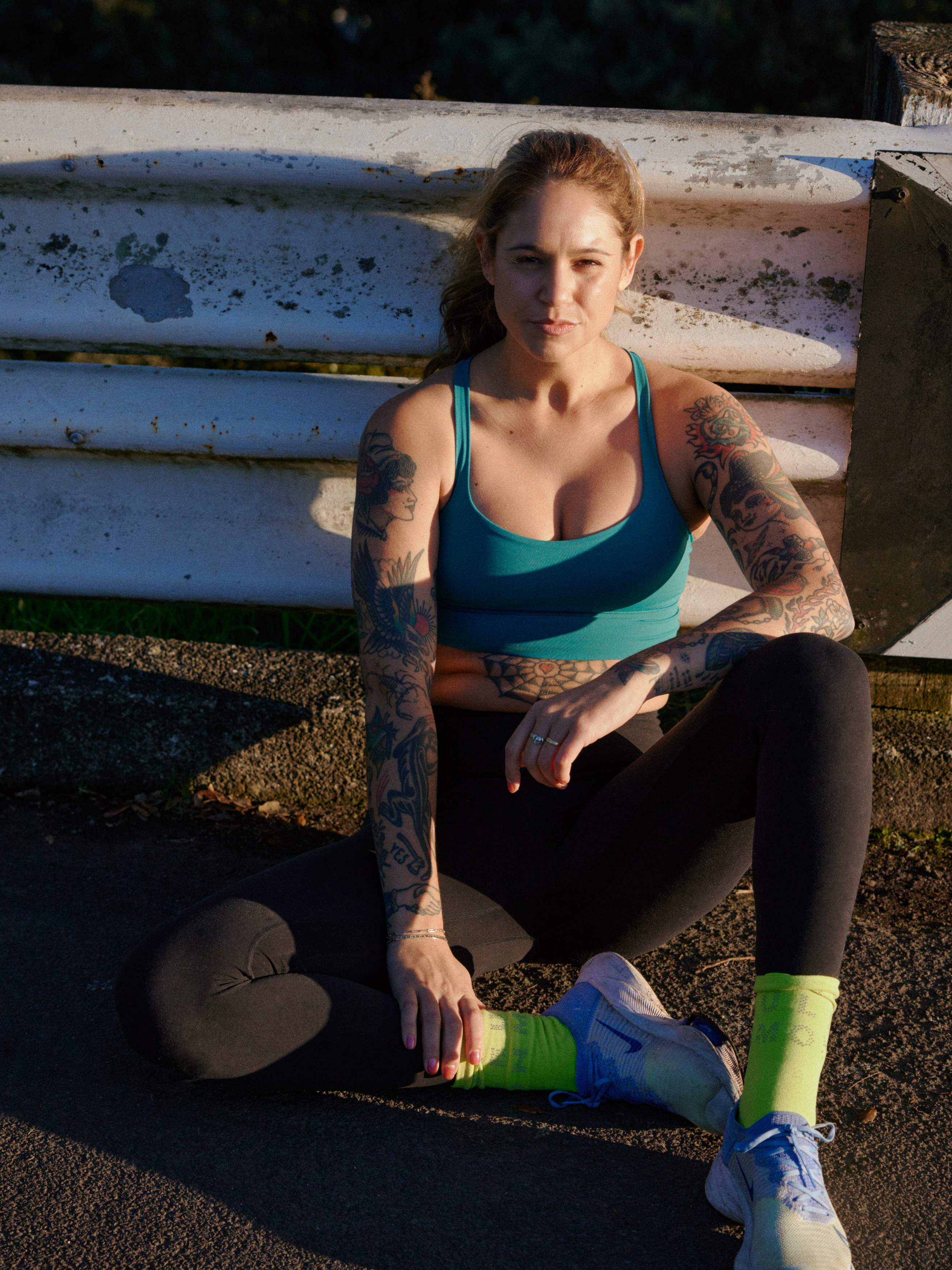Athletic woman with arm tattoos in workout clothes sitting outdoors on asphalt with a metal guard rail in the background.