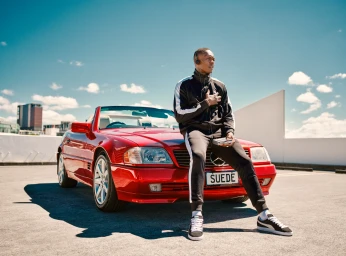 A Black man in a black tracksuit holding a portable cassette player while sitting on a red convertible car.