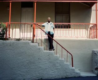 Black man in a light blue button-down shirt standing on outdoor stairs with a red railing by Reagen Butler