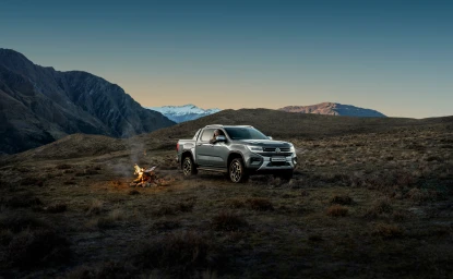 Silver pickup truck parked in a rugged mountain landscape at dusk, with a small campfire burning nearby.