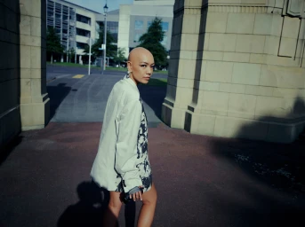 Bald woman wearing an oversized white blazer walking past neoclassical stone pillars on a city street