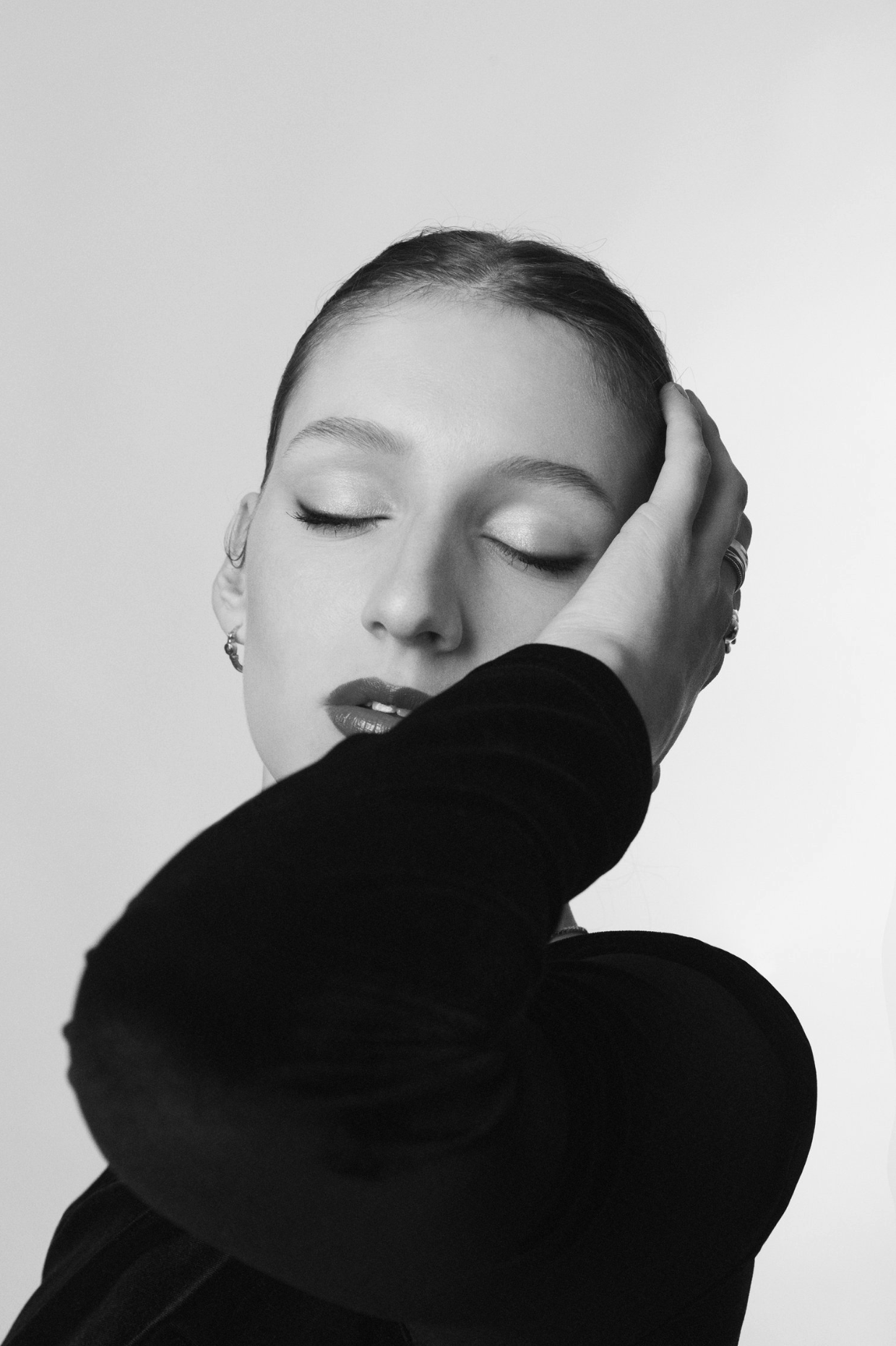 Monochrome portrait of a young woman with eyes closed, fair skin, red lips, and dark clothing, posing against a soft white background.
