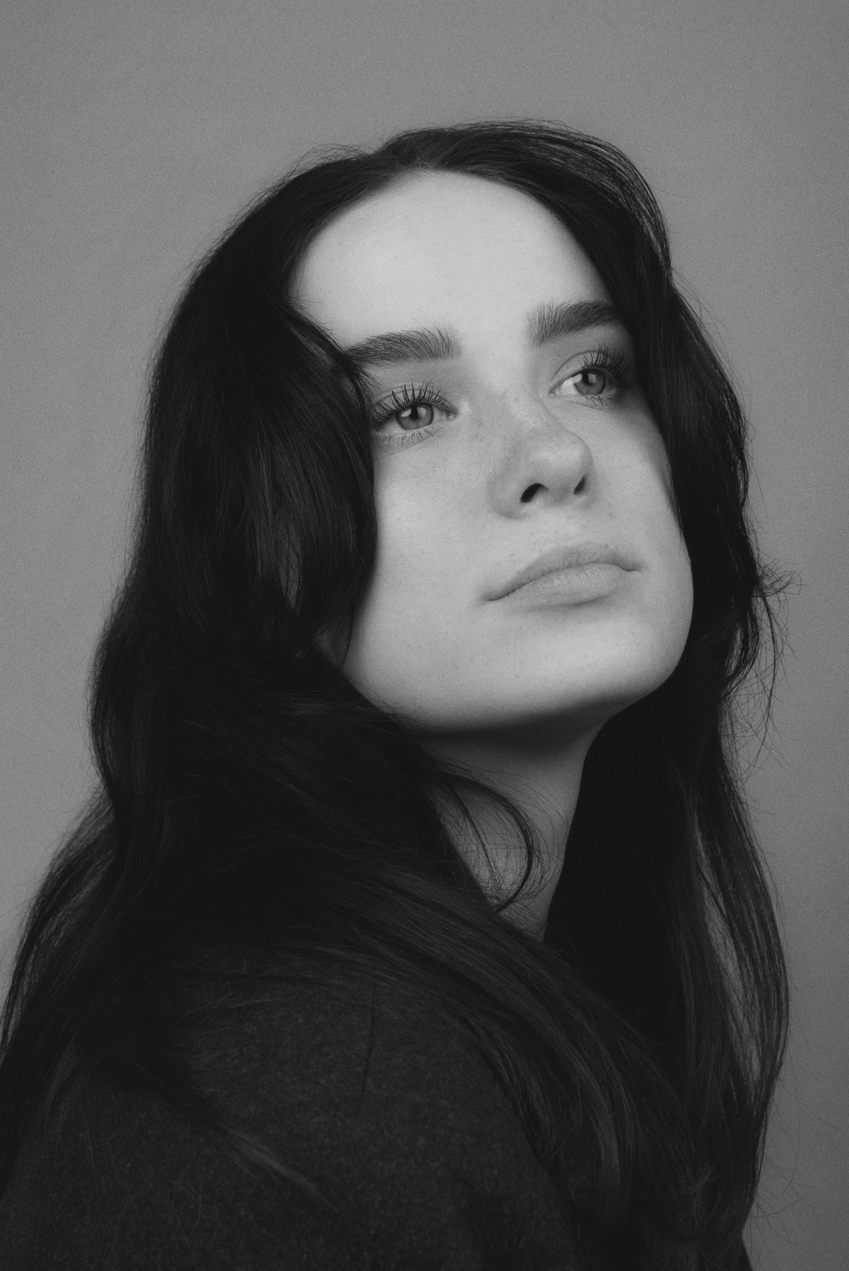 Black and white portrait of a young woman with long dark hair looking up against a solid gray background, captured in a studio setting.
