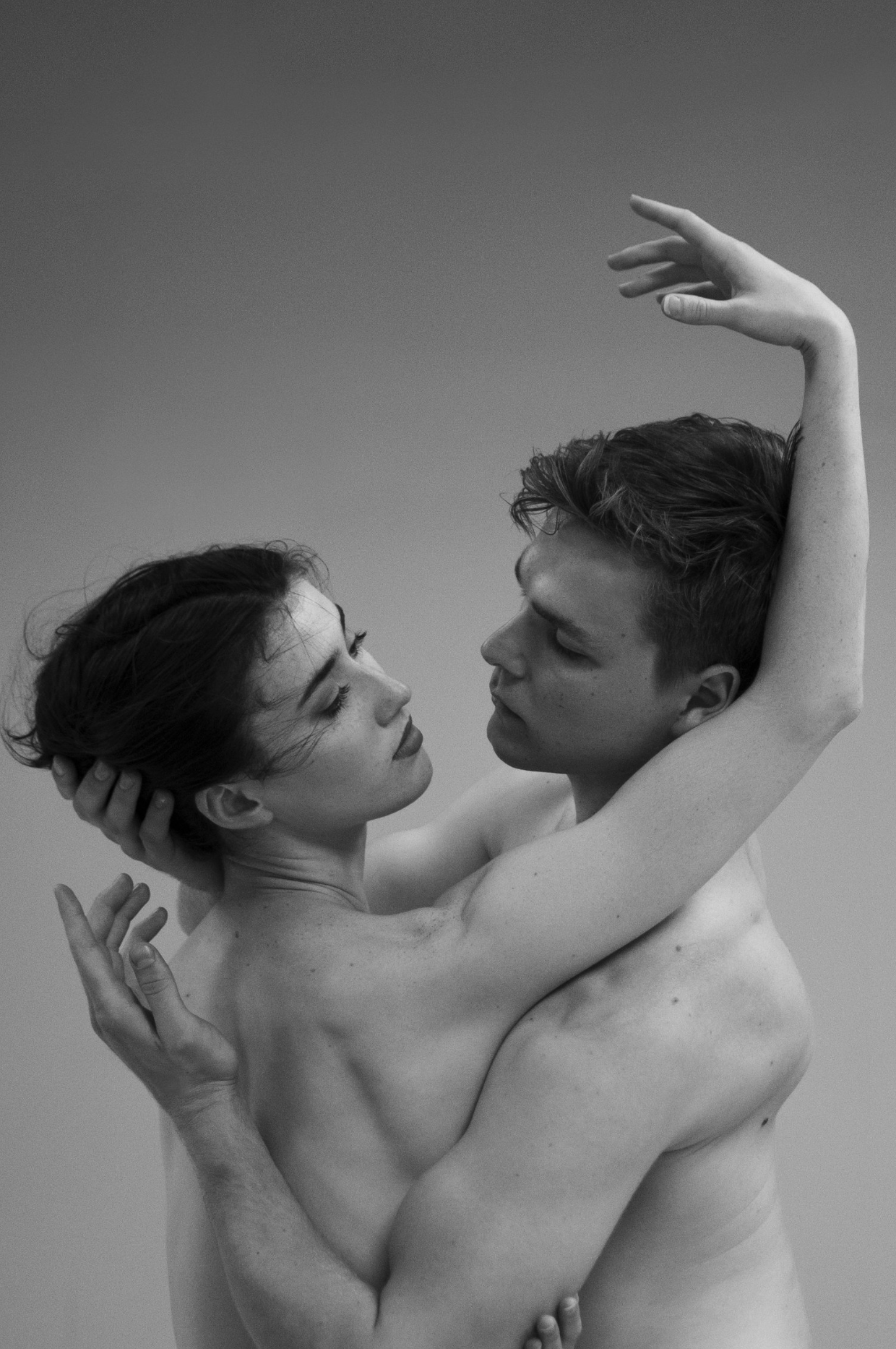Black and white studio shot of a nude couple embracing; the woman looks to the left and the man to the right with plain background.