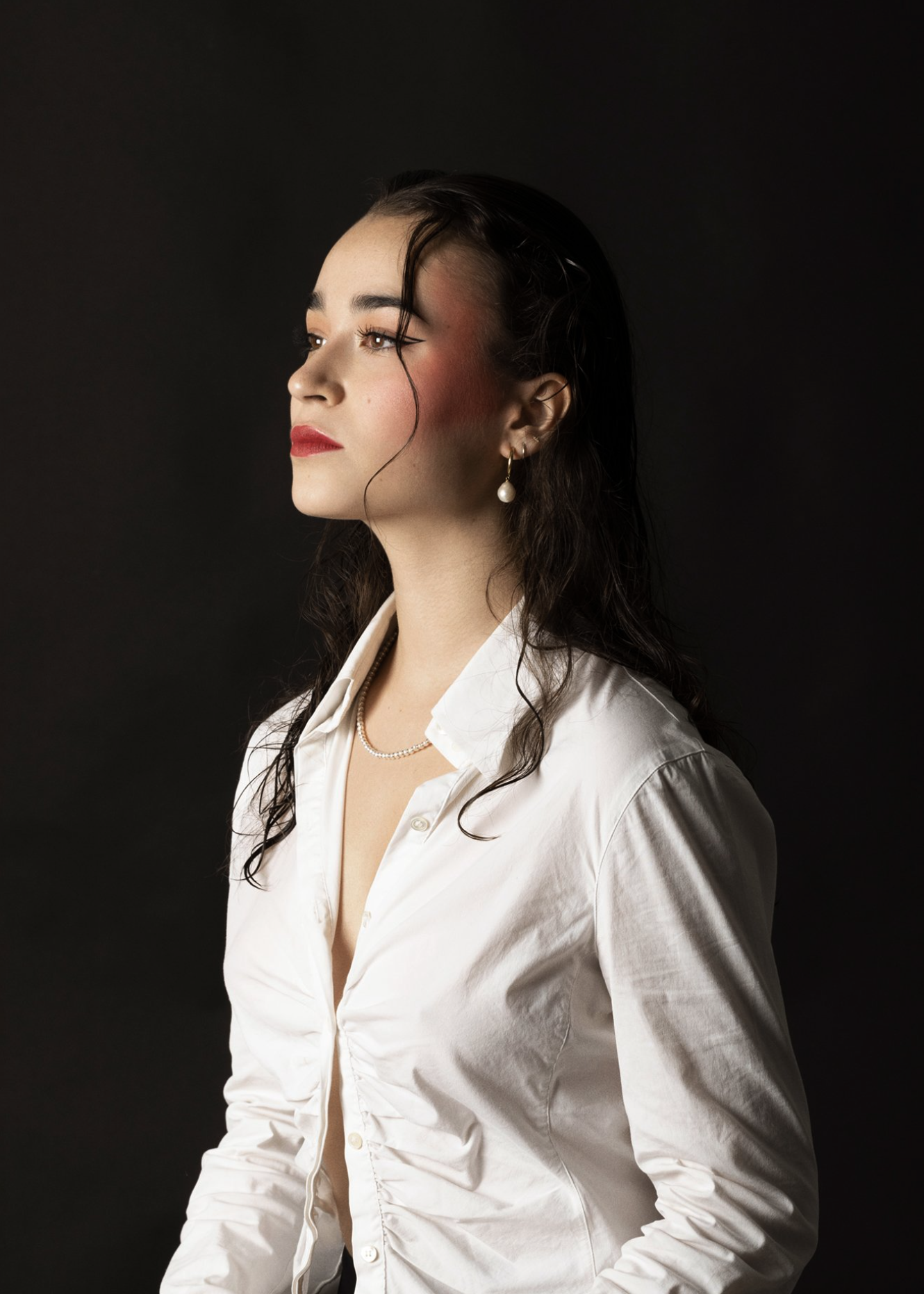 Portrait of a woman in a white shirt with wet hair, red lipstick, and heavy blush in a studio with a black background.