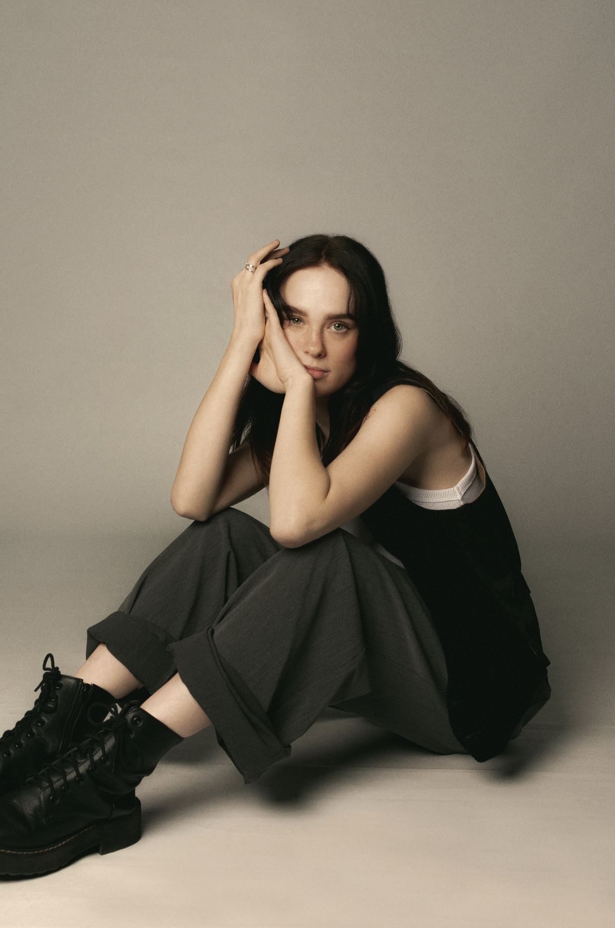 Young woman in a black tank top and grey pants sits on the floor with hands on her face and black boots in a studio setting.