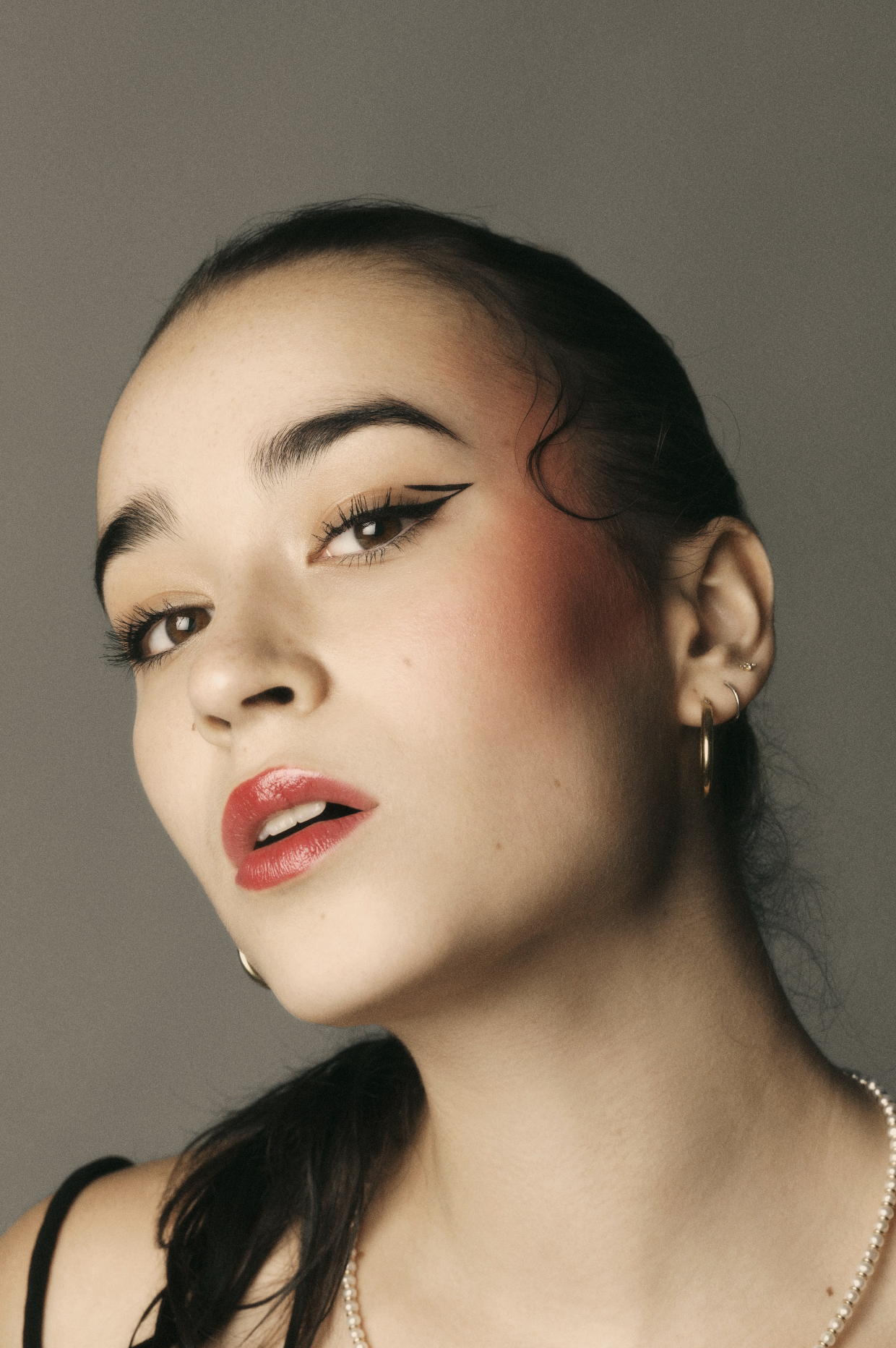 Close-up studio portrait of a young woman with bold eye makeup and glossy red lips against a grey background. Gold hoop earrings visible.