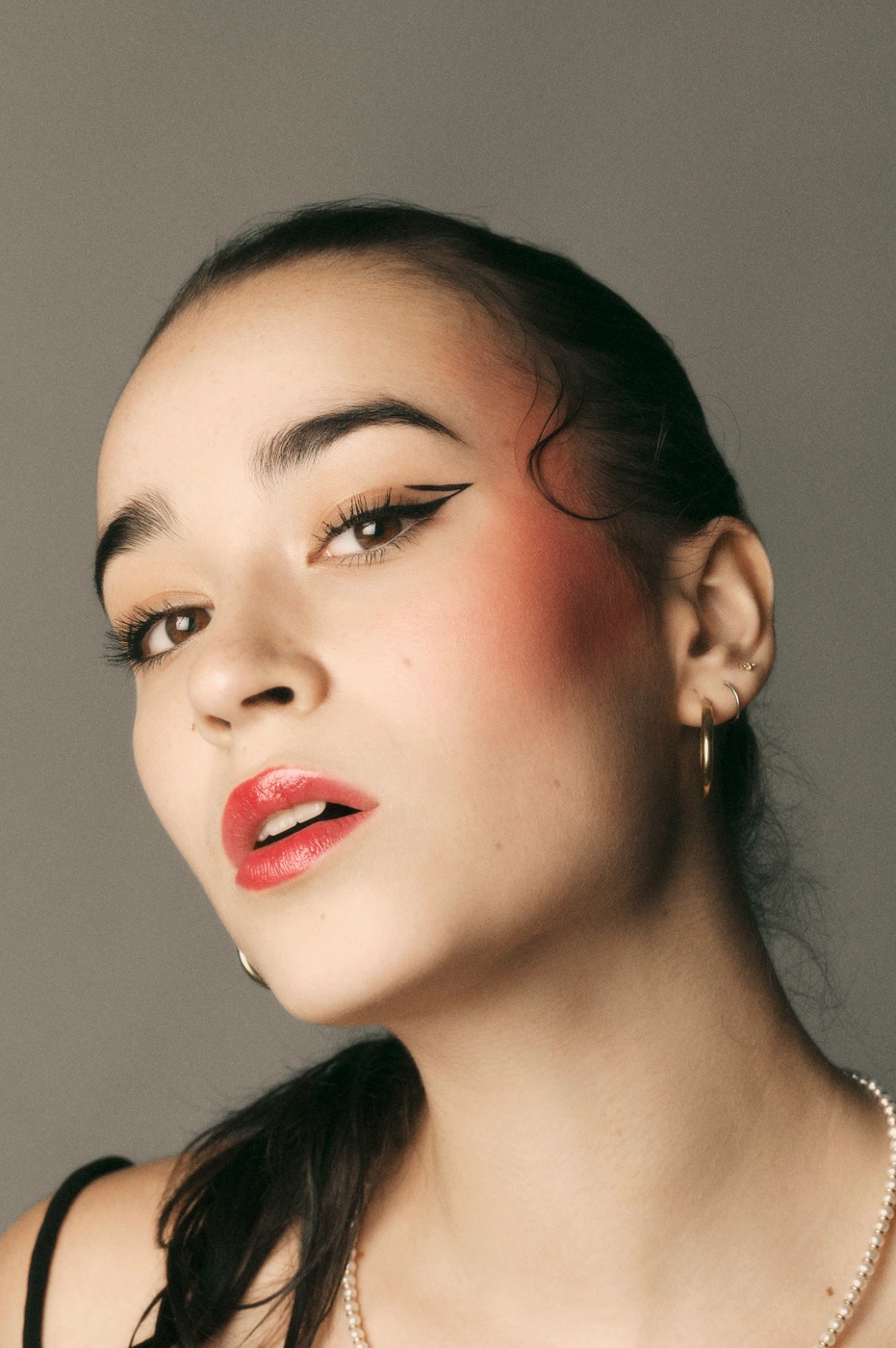 Close-up portrait of a young woman with graphic winged eyeliner, glossy red lips, and hair tied in a sleek ponytail against a grey backdrop.