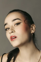 Close-up portrait of a young woman with graphic winged eyeliner, glossy red lips, and hair tied in a sleek ponytail against a grey backdrop.