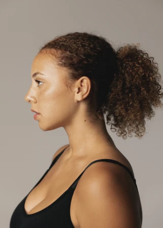 Profile portrait of a woman with curly brown hair in a ponytail wearing a black top against a plain grey background