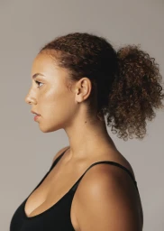 Profile portrait of a woman with curly brown hair in a ponytail wearing a black top against a plain grey background