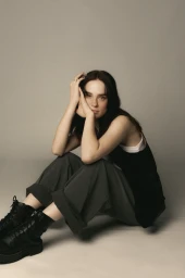 A woman with dark hair sitting on a plain grey seamless floor wearing dark trousers and black boots, posing with hands near her face.