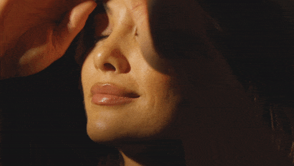 Close-up shot of an adult female face with a neutral expression, partially obscured by a hand, against a dark background.