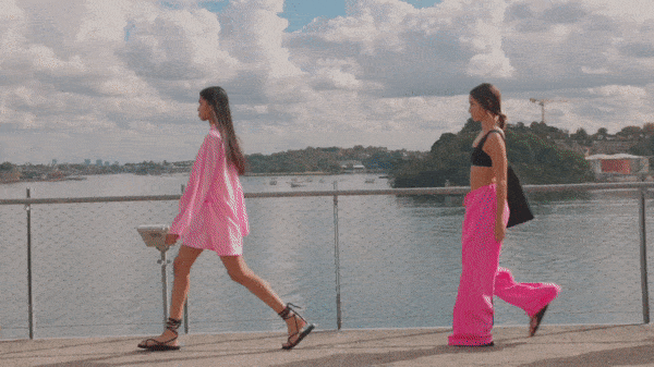 Two adult female fashion models in vibrant pink outfits walk on a waterfront walkway with a cityscape, blue sky, and clouds.
