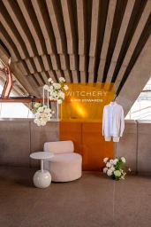 Fashion display featuring a white shirt on a mannequin, floral arrangements, modern armchair, and an orange branding panel under concrete architecture.
