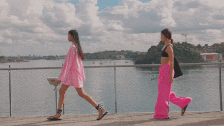 Two models walking across an outdoor bridge with water and clouds in the background