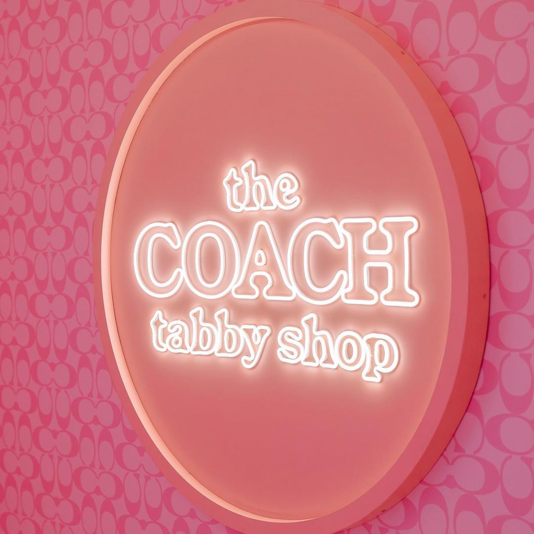 Illuminated round sign for The Coach Tabby Shop with a light pink background and a Coach logo patterned wall behind it.
